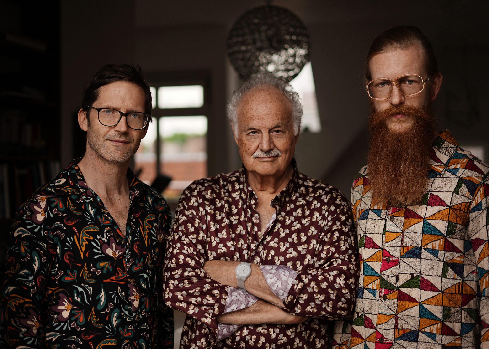 Drei Musiker des David Friedman Generations Trio stehen nebeneinander vor dunklem Hintergrund und tragen gemusterte Hemden.